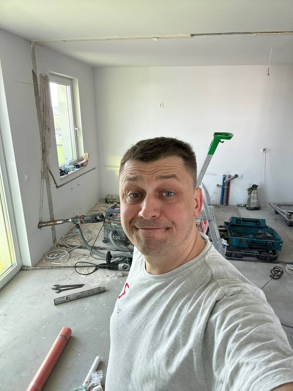 Mężczyzna na tle remontowanego pomieszczenia z narzędziami hydraulicznymi i instalacją przy oknie. Widoczny młot udarowy i drabina.