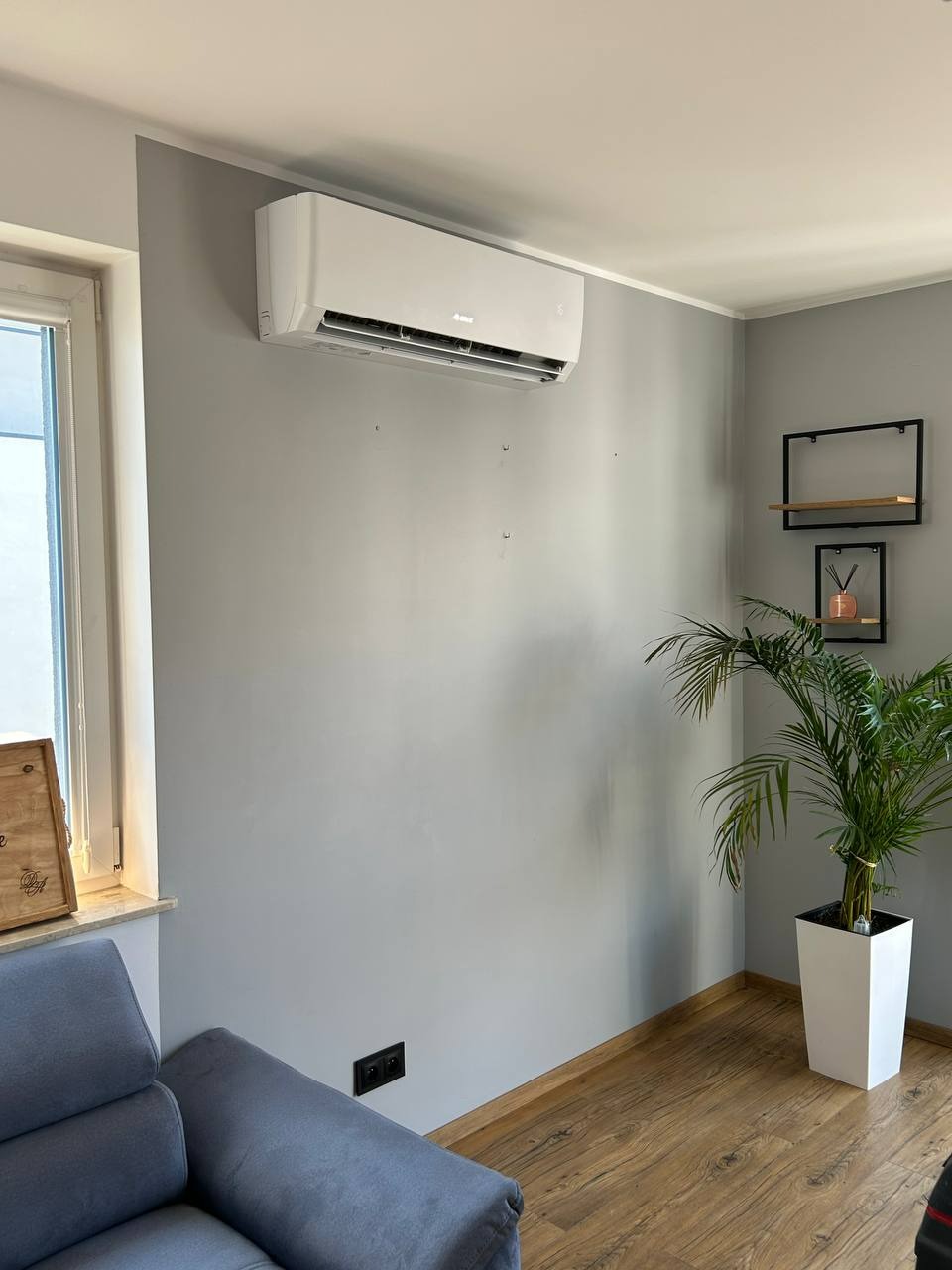 Biały klimatyzator na szarej ścianie w pokoju z drewnianą podłogą i rośliną doniczkową. Minimalistyczny design wnętrza.