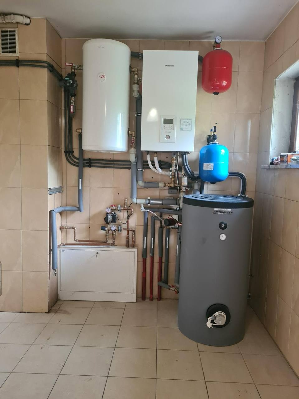 Kompletna kotłownia z szarym zasobnikiem wody, białym bojlerem i piecem gazowym Panasonic, zainstalowana w pomieszczeniu z beżowymi płytkami. Czerwony zbiornik wyrównawczy.