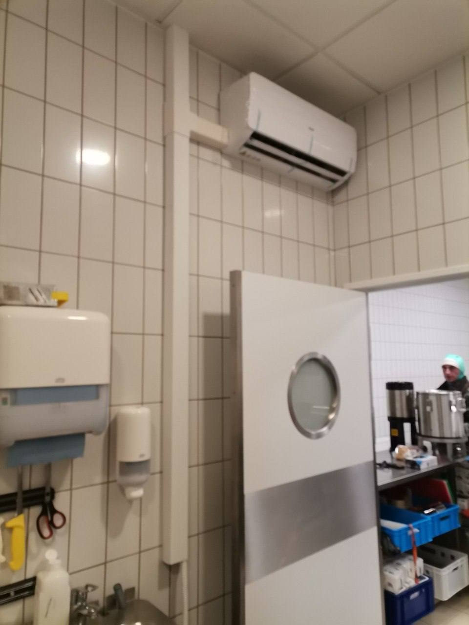 Biała klimatyzacja ścienna w pomieszczeniu wyłożonym kafelkami, z widocznym fragmentem kuchni przez okrągłe okienko w drzwiach.