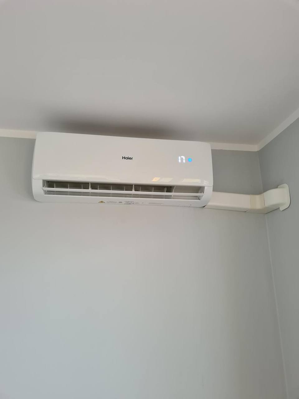 Biały klimatyzator Haier zamontowany na ścianie, z widocznymi przewodami w białej maskownicy. Na wyświetlaczu widoczna temperatura 17 stopni.