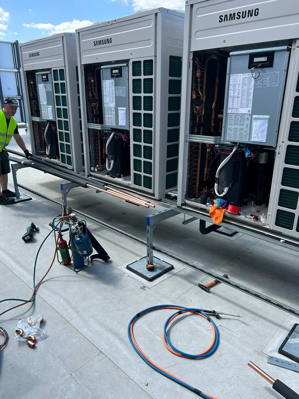 Montaż instalacji klimatyzacji Samsung na dachu budynku. Widoczne narzędzia, rury miedziane i pracownik w kamizelce. Detaliczny widok na etap instalacji.