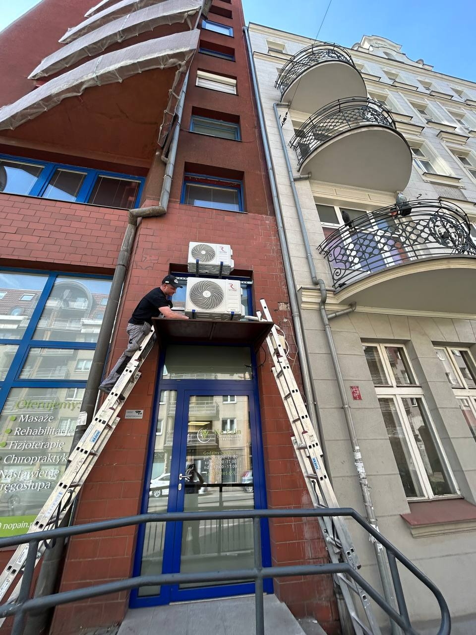 Mężczyzna na drabinie montuje klimatyzatory na elewacji budynku z czerwonej cegły, obok budynku z balkonami. Widok z dołu na instalację klimatyzacji.