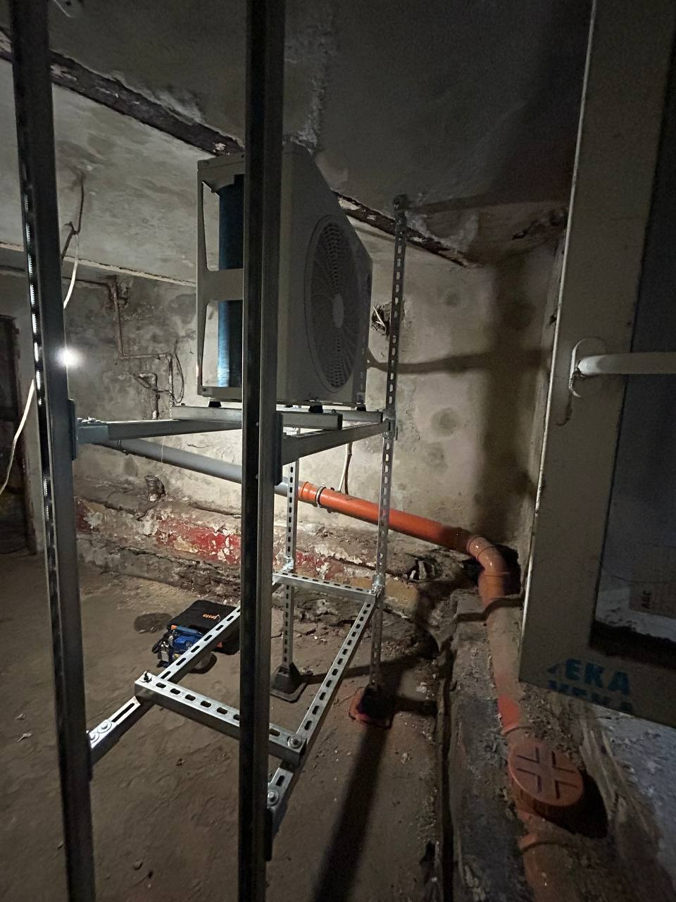 Montaż klimatyzacji w piwnicy: jednostka zewnętrzna na stalowej konstrukcji wsporczej, widoczne rury odprowadzające skropliny. Surowy stan pomieszczenia.