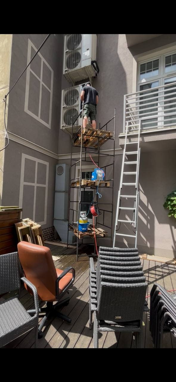Mężczyzna na rusztowaniu z palet montuje klimatyzator na elewacji budynku. Widoczne dwa klimatyzatory, drabina i narzędzia na podeście rusztowania.