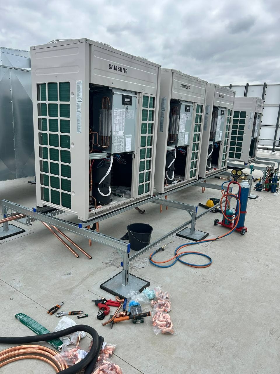 Rząd jednostek klimatyzacyjnych Samsung na dachu, instalacja w toku. Widoczne narzędzia, rury miedziane i butla z gazem. Szare niebo w tle.