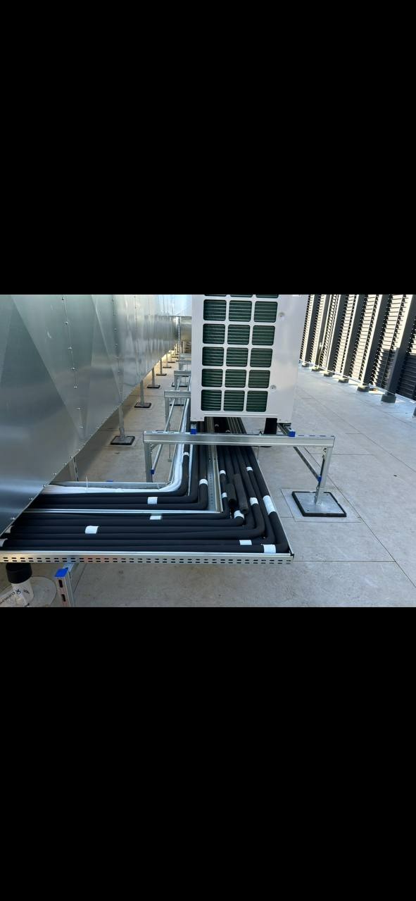 Instalacja rur w izolacji termicznej na zewnątrz budynku, poprowadzona w korytach kablowych. Widoczny agregat klimatyzacji i elementy konstrukcyjne dachu.