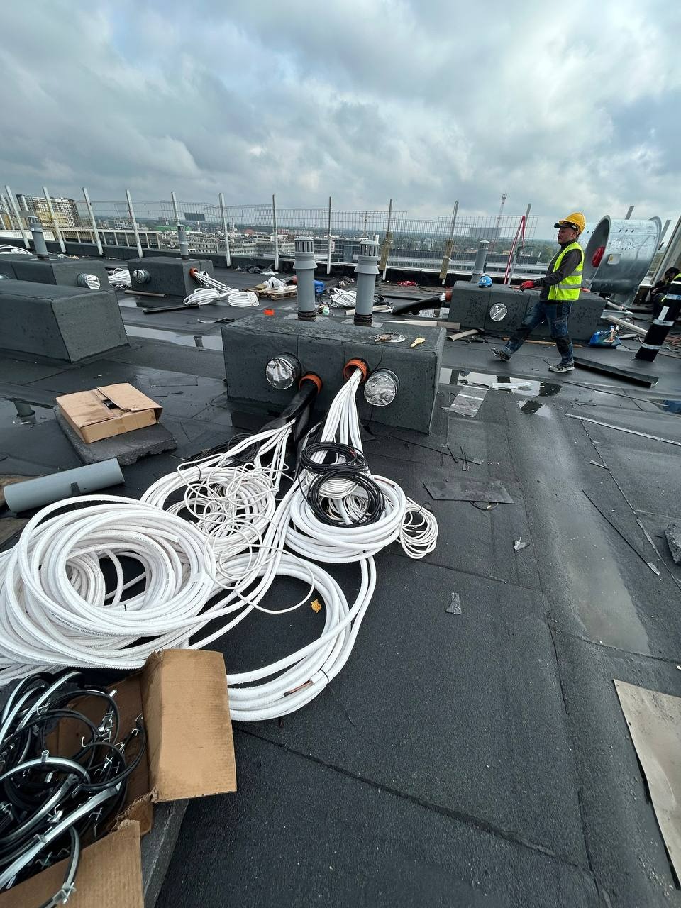 Instalacja przewodów wentylacyjnych na dachu budynku, pracownik w kasku w tle. Widoczne elementy wentylacji i przewody w rolkach. Pochmurne niebo.