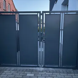 Nowoczesna, grafitowa brama wjazdowa z metalowymi panelami, widoczna na tle brukowanej nawierzchni i budynków. Minimalistyczny design, zamknięta pozycja.