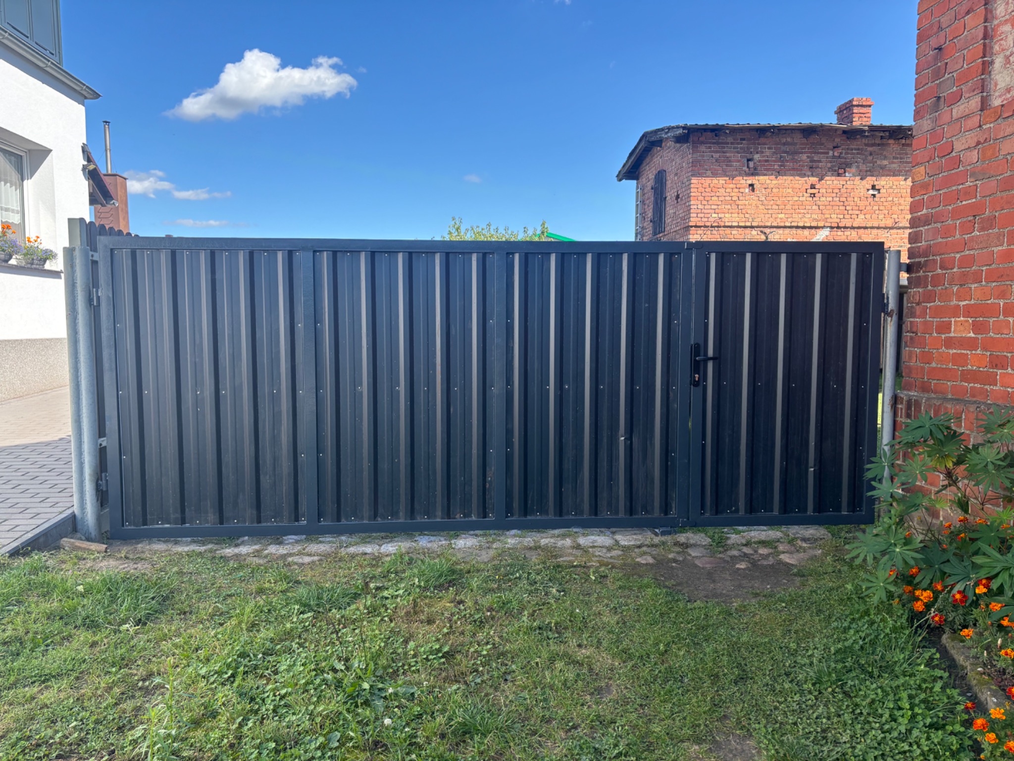 Ciemnoszara brama wjazdowa z metalowych paneli pionowych, z czarną klamką, na tle zielonego trawnika i zabudowań z czerwonej cegły, w słoneczny dzień.