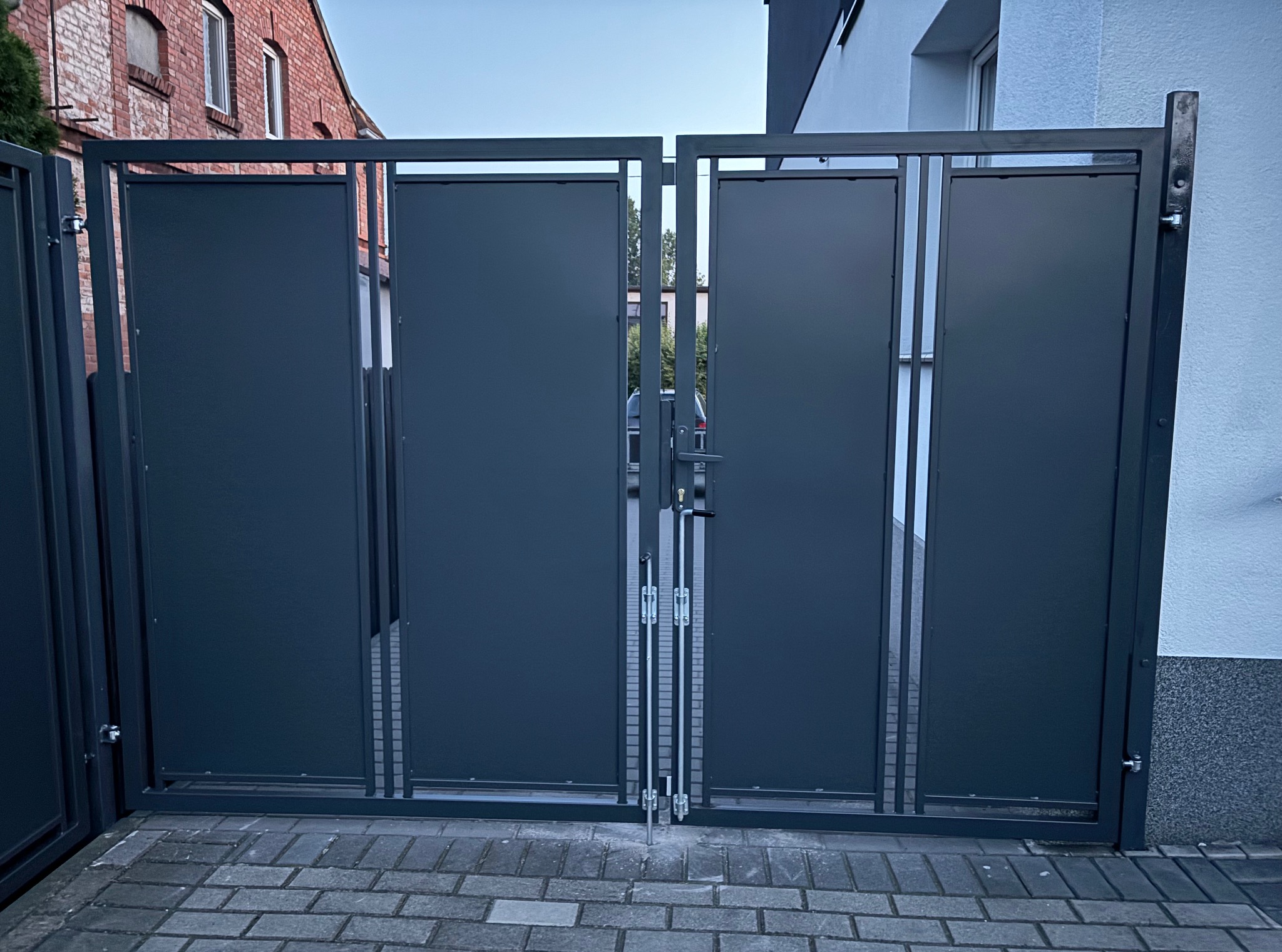 Nowoczesna, grafitowa brama wjazdowa z metalowymi panelami, widoczna na tle brukowanej nawierzchni i budynków. Minimalistyczny design, zamknięta pozycja.
