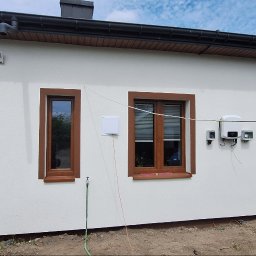 Damseb - Biała elewacja domu z brązowymi detalami okien i narożników. Widoczne okna z żaluzjami, instalacja solarna i fragment osoby w niebieskim kombinezonie.