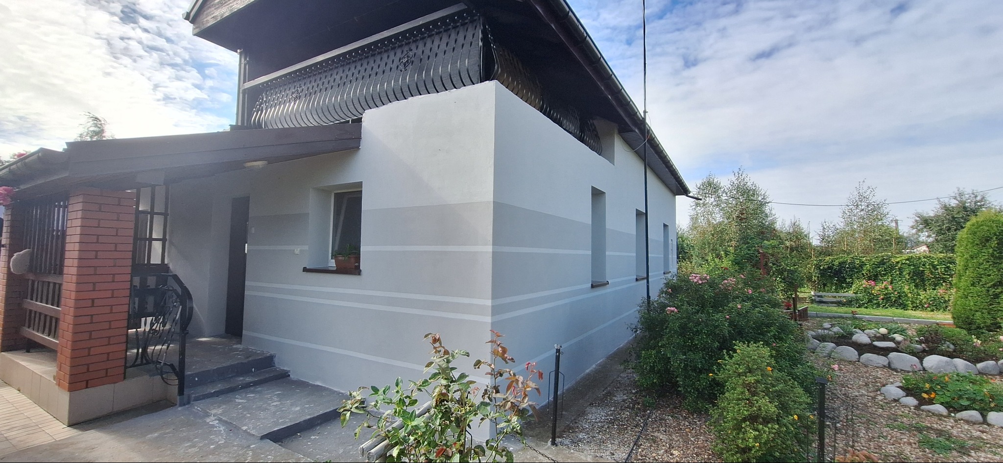 Odnowiona elewacja domu z szarymi pasami, widok z boku, z elementami ogrodu. Balustrada balkonu z metaloplastyki. Wejście z ceglanym filarem.