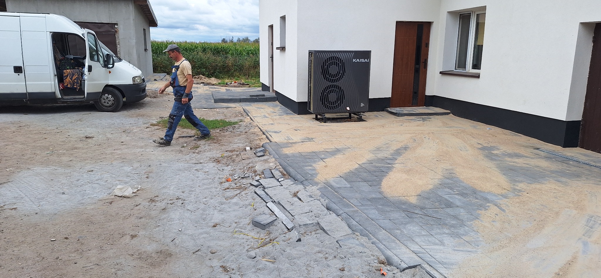 Układanie szarej kostki brukowej na podwórku domu, widoczny pracownik w kombinezonie i bus, częściowo ułożona kostka, piasek na kostce, budynek w tle.