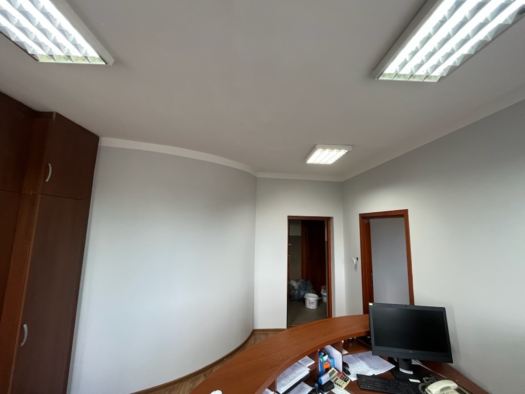 Jasne biuro po malowaniu: szare ściany, drewniana zabudowa, widoczne drzwi i oświetlenie sufitowe. Minimalistyczny design z elementami wyposażenia biurowego.