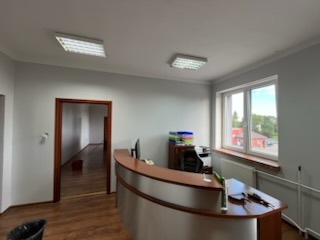 Jasne biuro po remoncie z drewnianą podłogą i recepcją. Widok na puste pomieszczenie przez otwarte drzwi. Nowoczesne oświetlenie sufitowe.