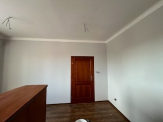 Szare ściany z białym gzymsem, drewniane drzwi w odnowionym pokoju. Widoczne wystające kable. Fragment komody w rogu. Minimalistyczne wykończenie wnętrza.