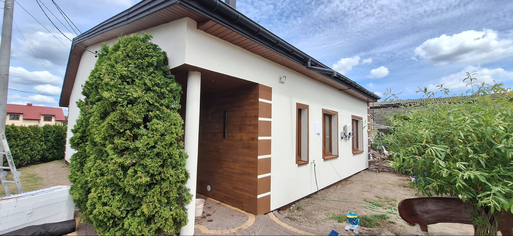 Wykończony budynek z drewnianą elewacją, kolumną i oknami w brązowych ramach. Widok na fragment ogrodu i błękitne niebo z chmurami.