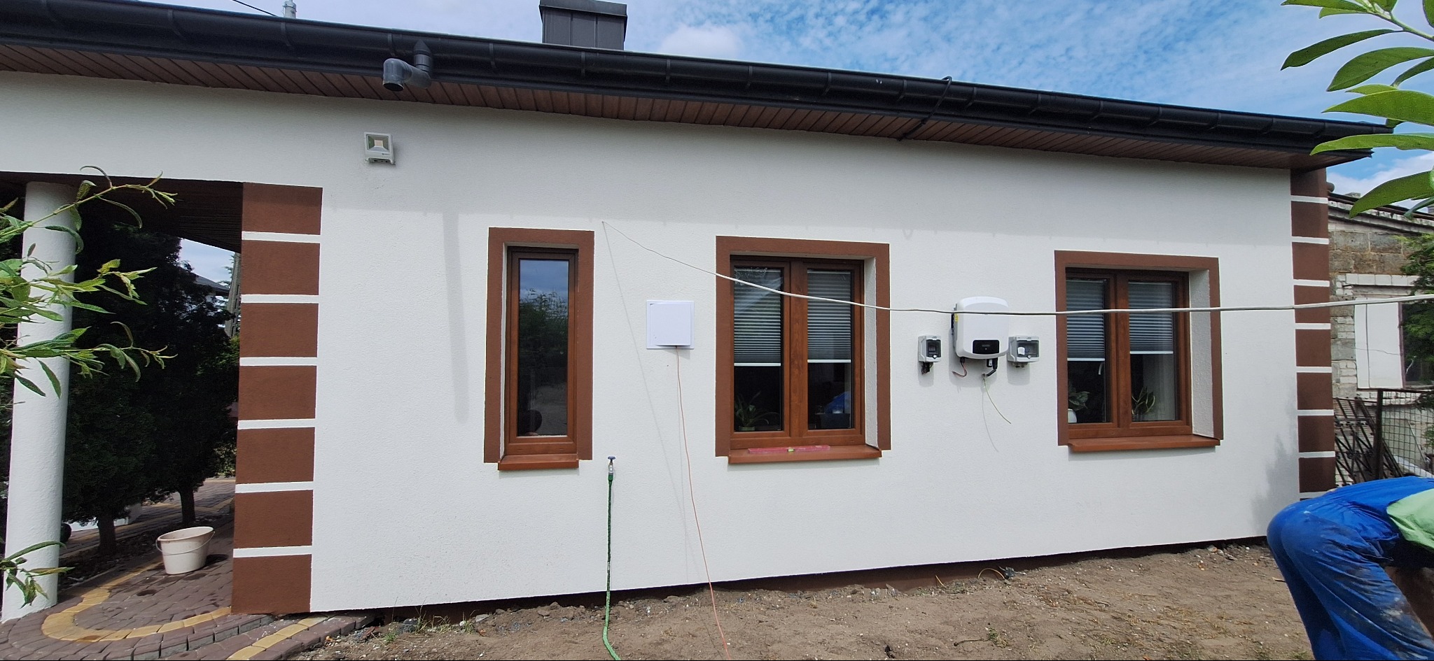 Biała elewacja domu z brązowymi detalami okien i narożników. Widoczne okna z żaluzjami, instalacja solarna i fragment osoby w niebieskim kombinezonie.