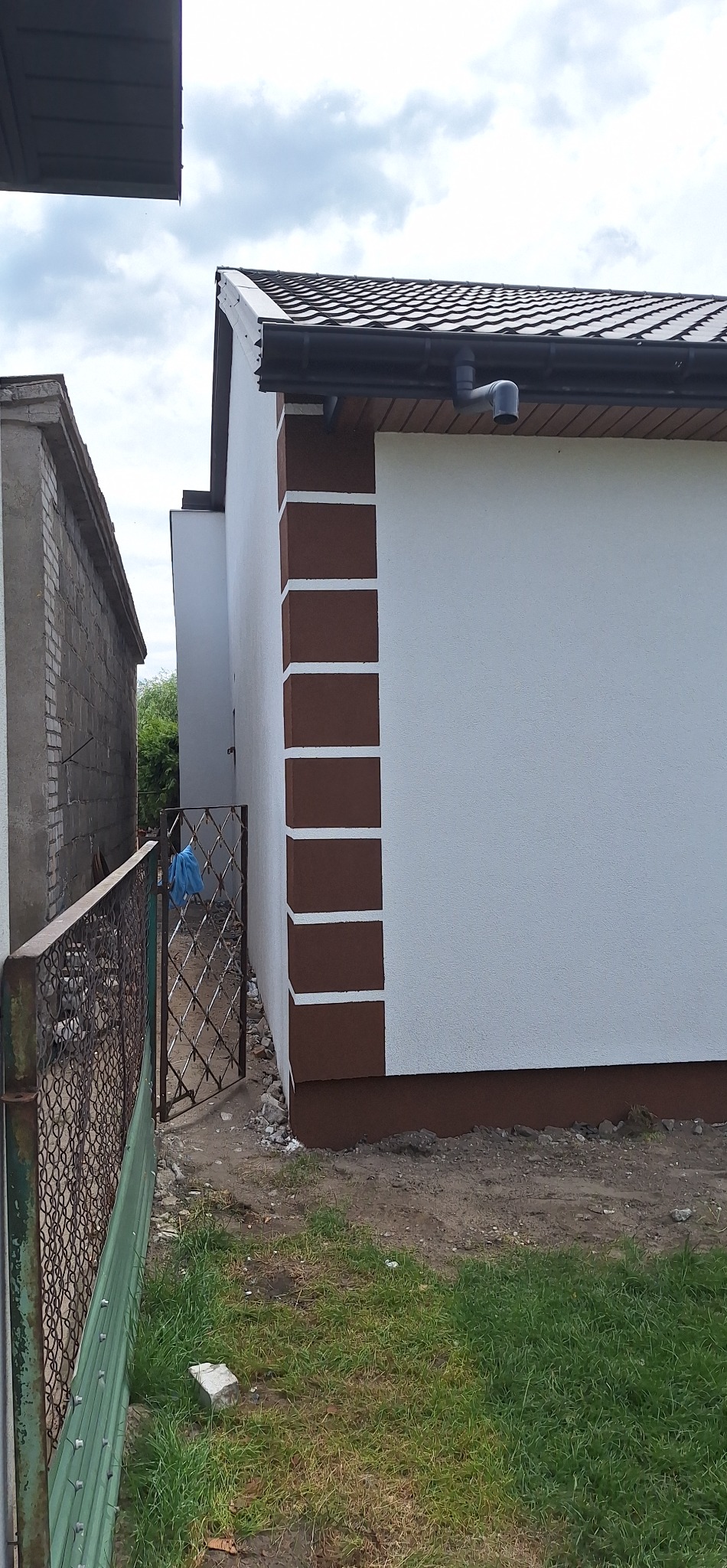 Ściana domu po remoncie z dekoracyjnymi, brązowymi elementami. Widoczny fragment dachu z rynną i brama wjazdowa.