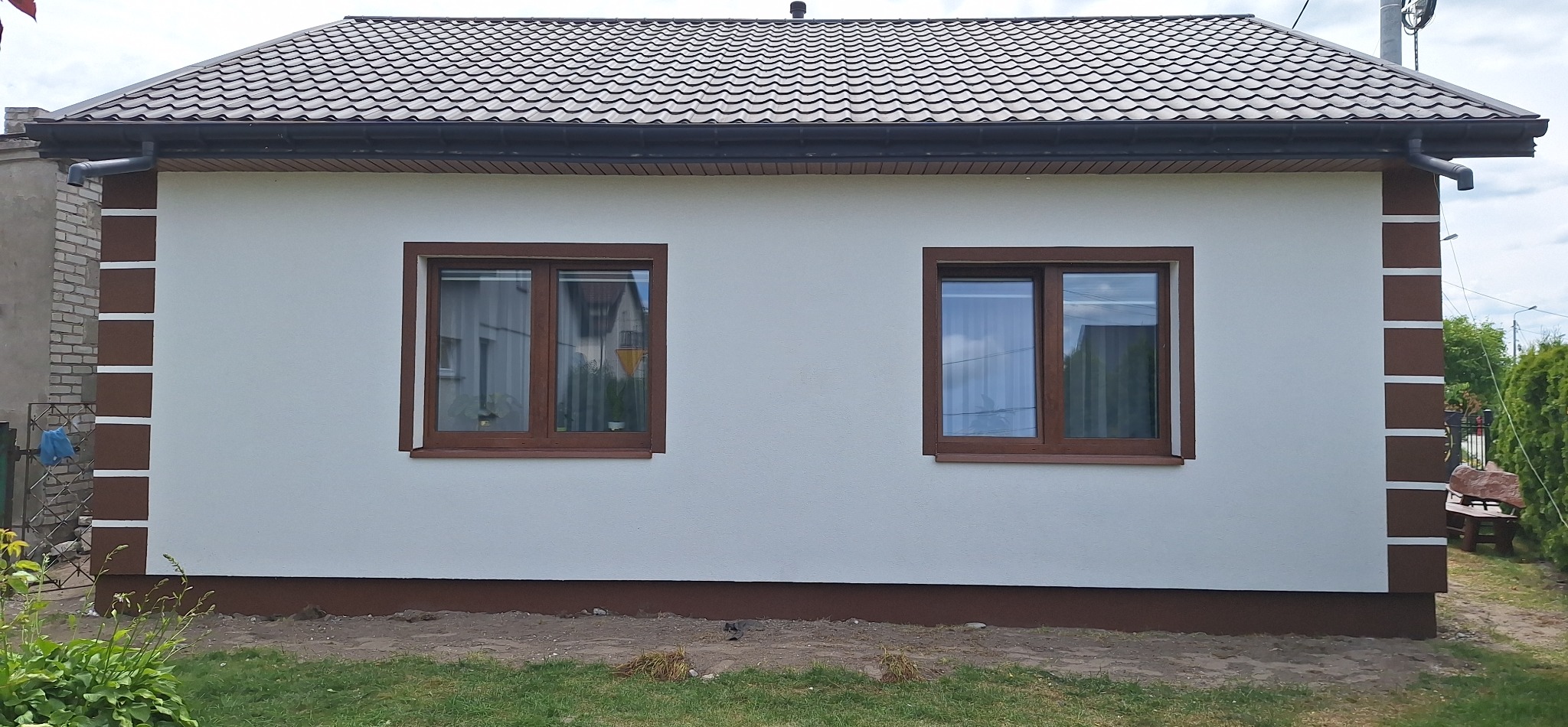 Elewacja domu jednorodzinnego po remoncie, z brązowymi akcentami na rogach i pod oknami. Widoczne dwa okna z brązowymi ramami i brązowy dach.