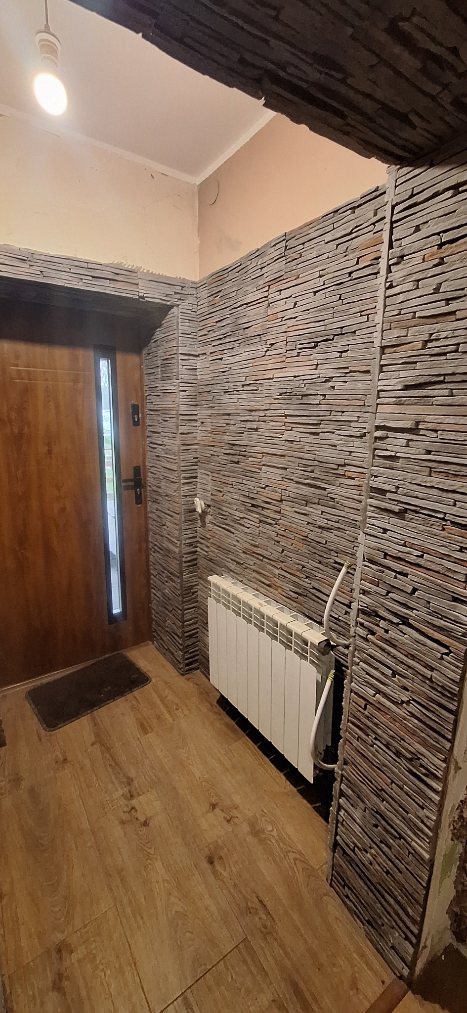 Wykończone wnętrze przedpokoju z dekoracyjną okładziną kamienną na ścianach, drewnianą podłogą i grzejnikiem. Widoczne drzwi wejściowe z szybą.