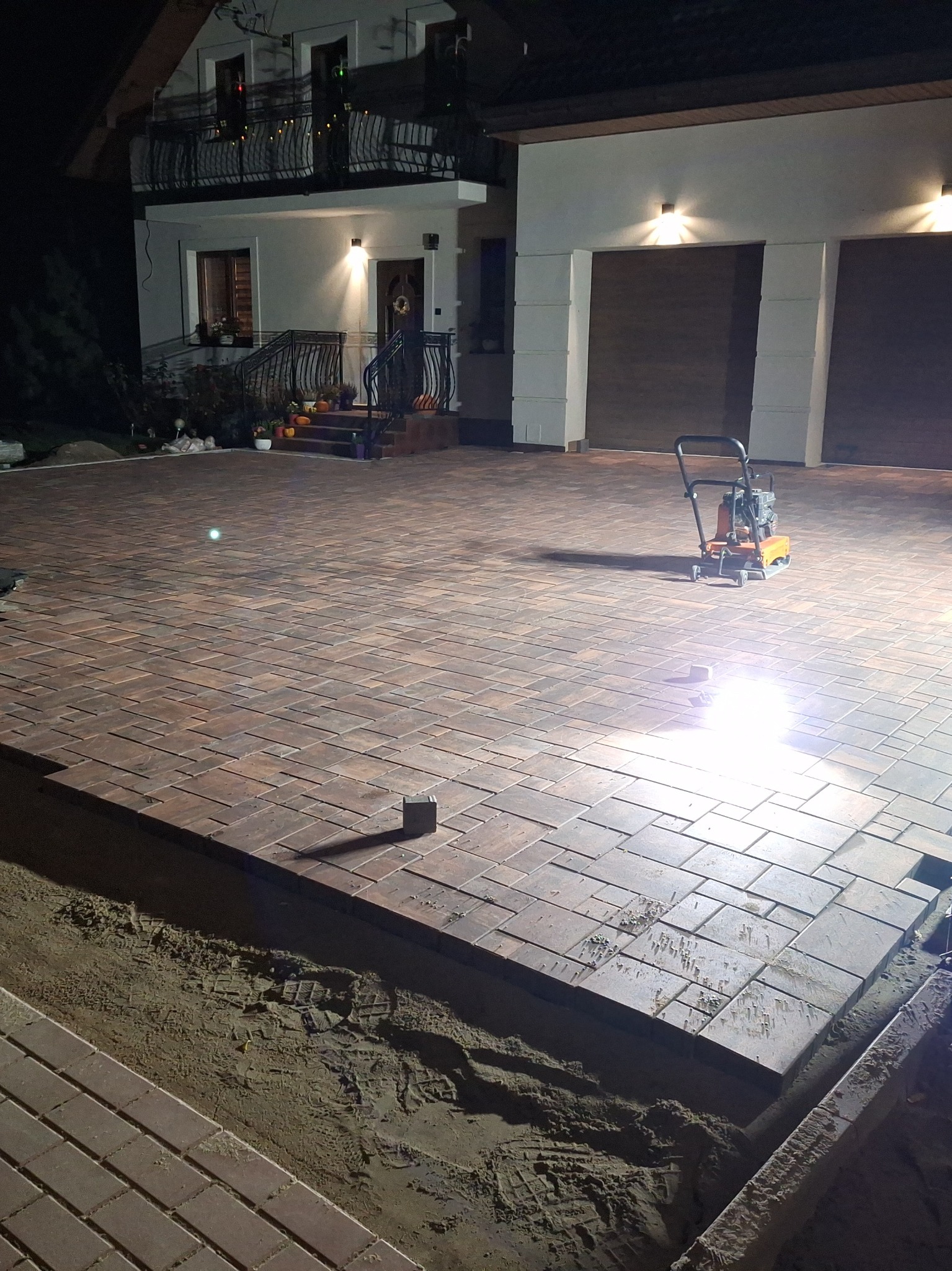 Układanie kostki brukowej na posesji domu jednorodzinnego nocą, widoczna ubijarka w trakcie pracy, częściowo ułożona kostka, przygotowane podłoże.
