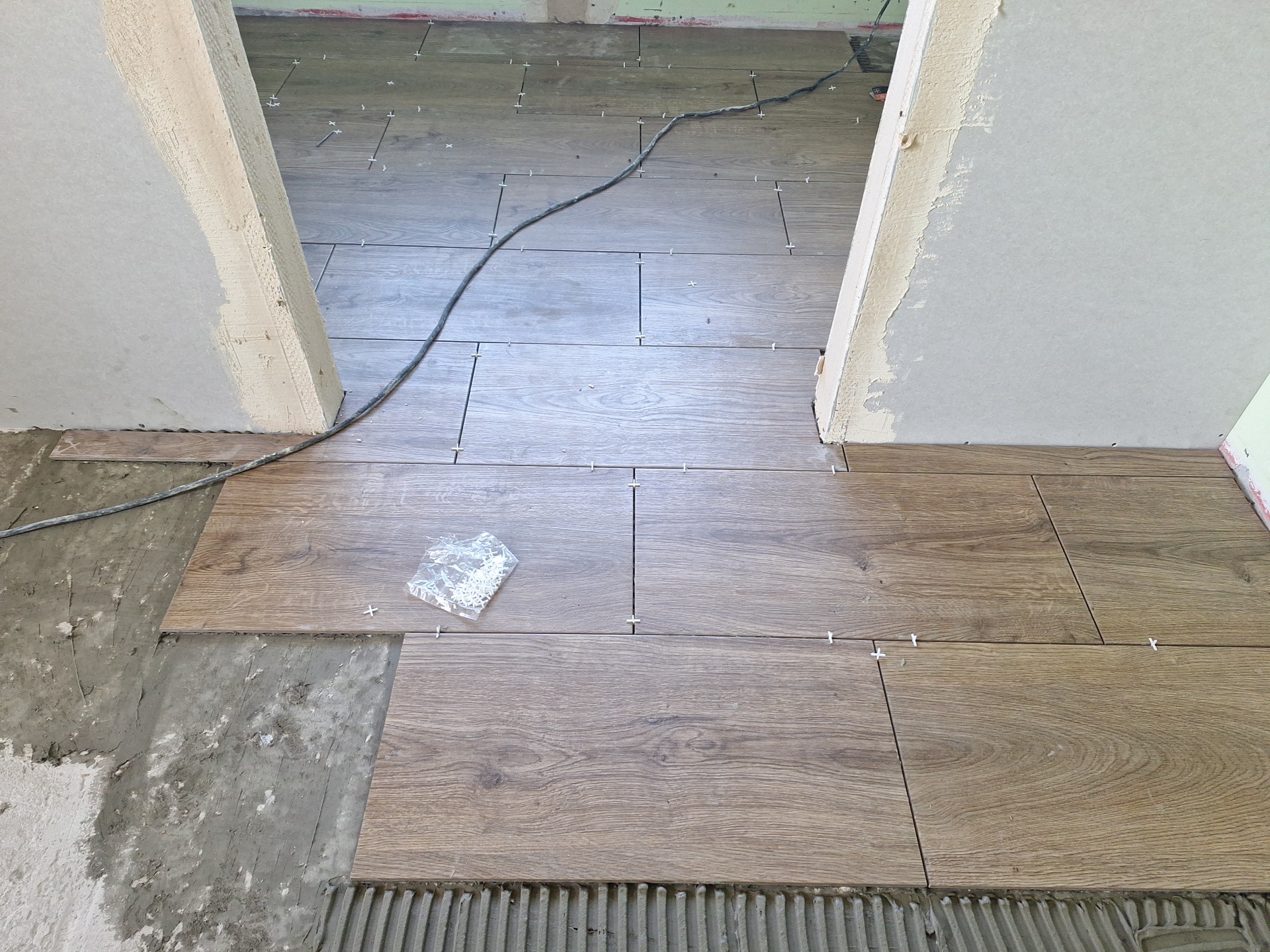 Układanie drewnopodobnych płytek podłogowych w przejściu, widoczne krzyżyki dystansowe i paczka z krzyżykami. Prace glazurnicze w Szreńsku.