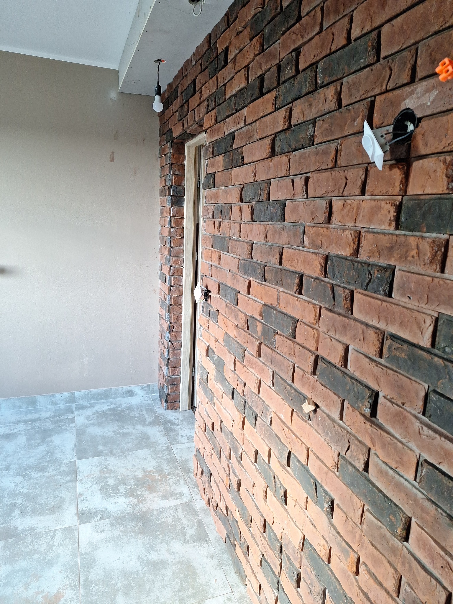 Ściana wykończona czerwoną cegłą klinkierową w pomieszczeniu z szarą podłogą z płytek i widoczną instalacją elektryczną. Widoczny fragment ościeżnicy drzwi.