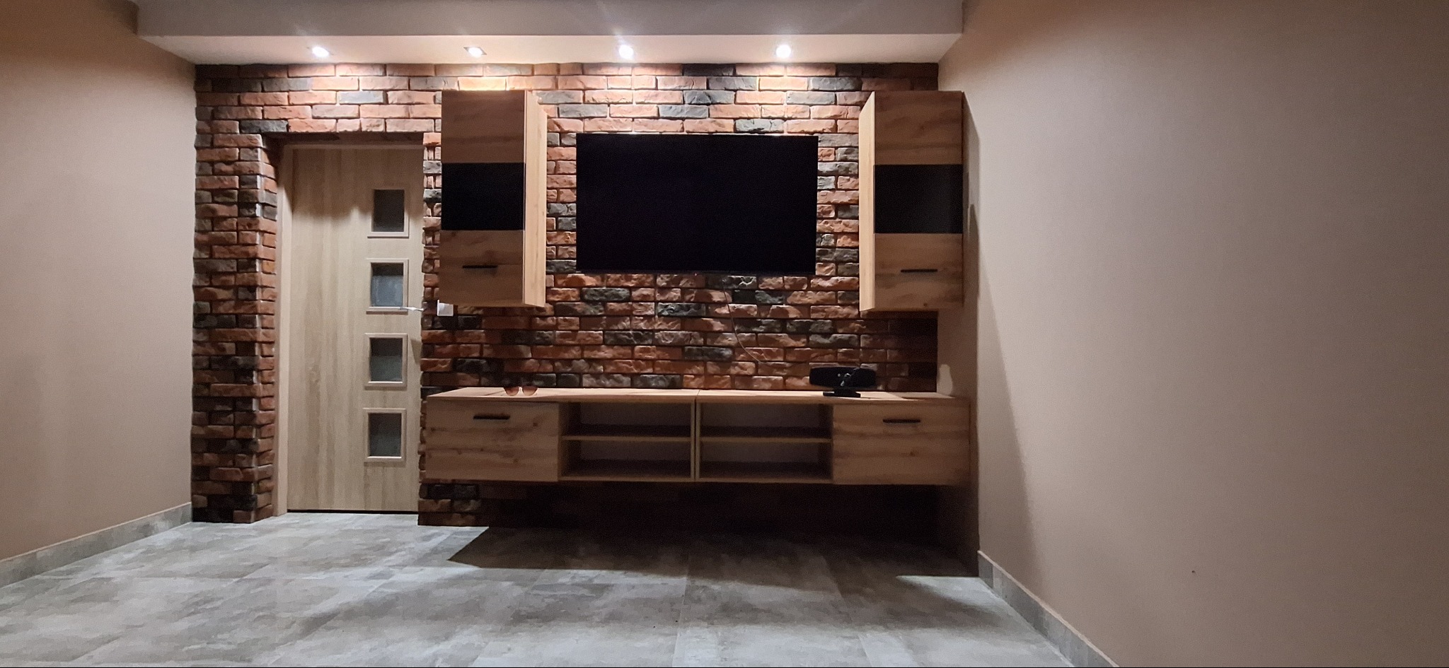 Wykończony salon z ceglaną ścianą, drewnianymi szafkami RTV i drzwiami z przeszkleniami. Oświetlenie punktowe podkreśla strukturę cegły.