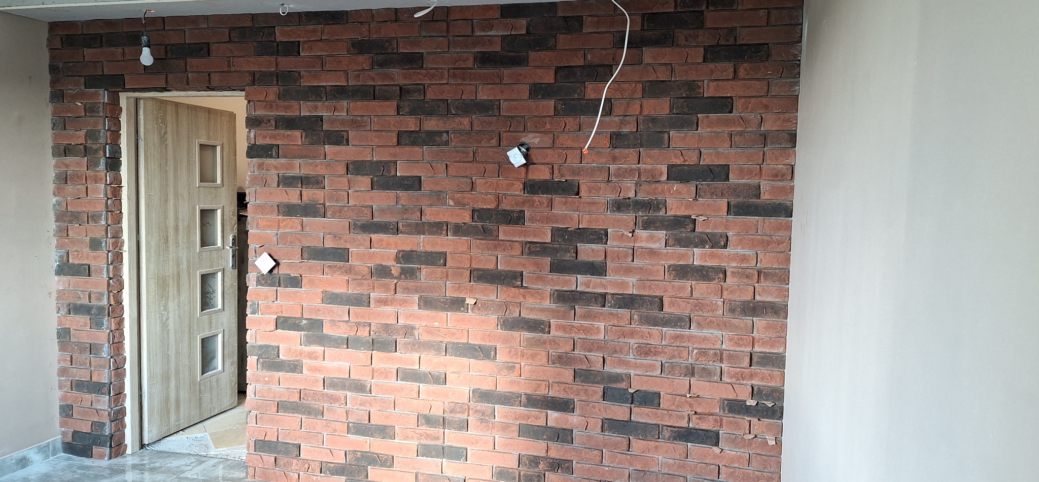 Ściana z cegły klinkierowej w odcieniach czerwieni i czerni, z widocznym fragmentem ościeżnicy i drzwi. Fragment ściany obok cegieł jest w trakcie przygotowania do malowania.