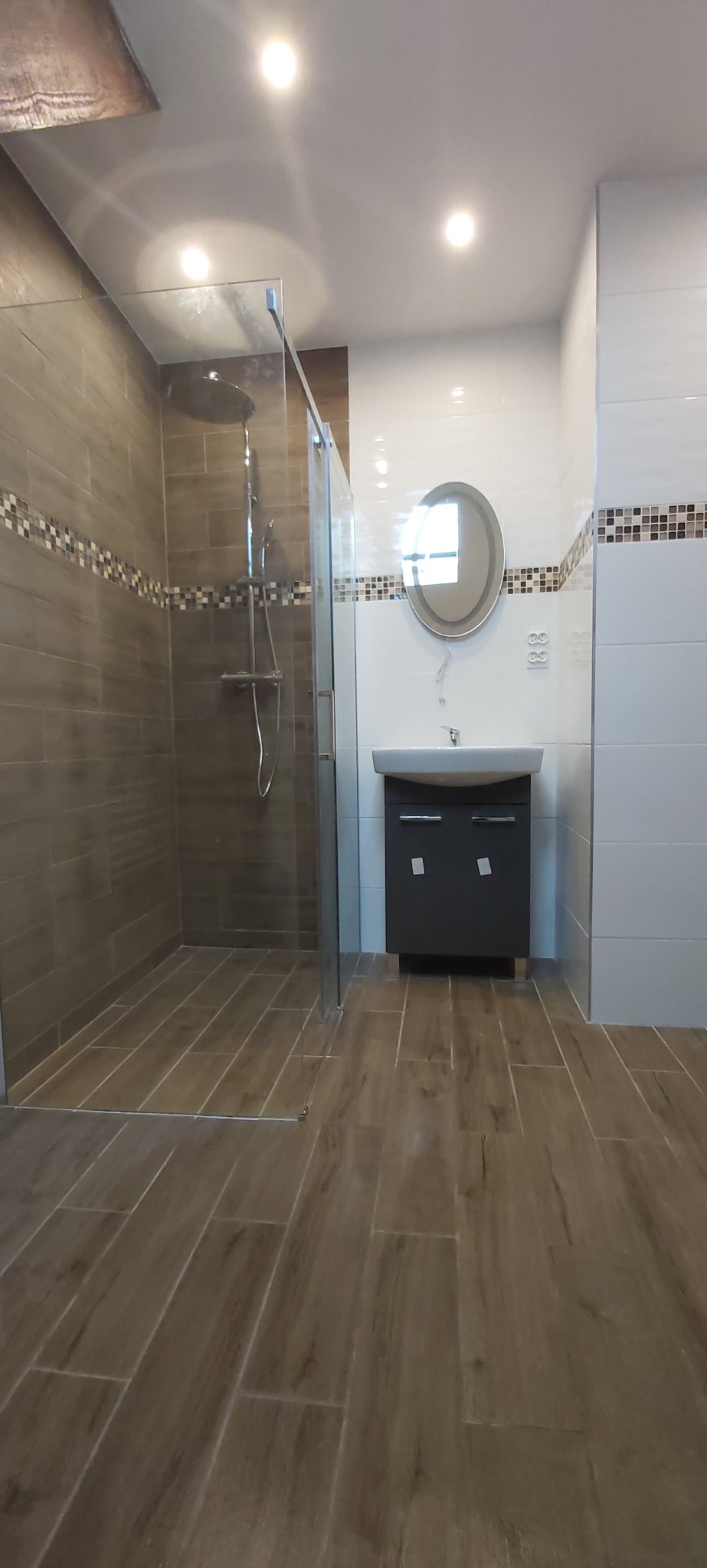 Nowoczesna łazienka po remoncie: kabina prysznicowa typu walk-in z brązowymi płytkami imitującymi drewno, umywalka z szafką i okrągłe lustro.