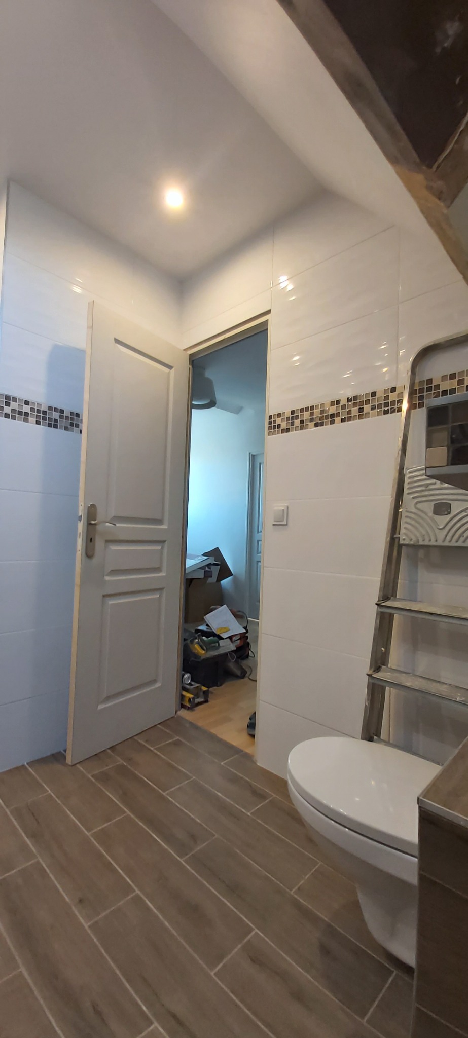 Remont łazienki w trakcie: białe płytki z mozaiką, brązowe płytki podłogowe imitujące drewno, drabina i częściowo widoczna toaleta, otwarte drzwi do innego pomieszczenia.