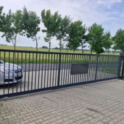 Nowoczesna, przesuwna brama wjazdowa z metalowymi sztachetami na posesji, z widocznym fragmentem samochodu i zielonym tłem drzew i pola. Brama w kolorze grafitowym.