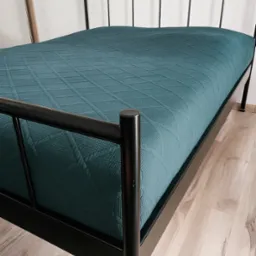 Czarne metalowe łóżko z turkusową narzutą o geometrycznym wzorze, na jasnej podłodze. Widoczne odbicie w lustrze i fragment białej ściany. Minimalistyczny design.