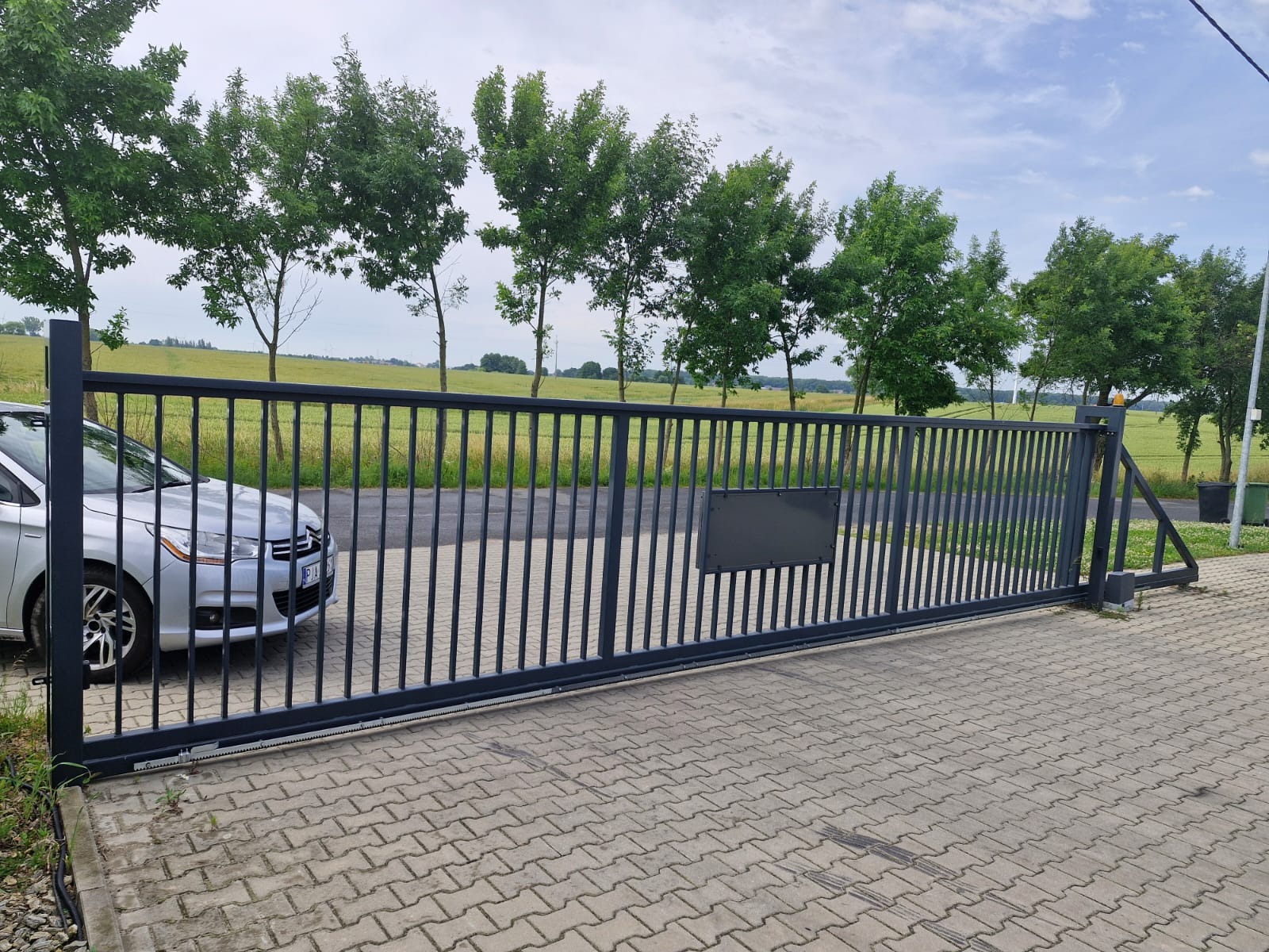 Nowoczesna, przesuwna brama wjazdowa z metalowymi sztachetami na posesji, z widocznym fragmentem samochodu i zielonym tłem drzew i pola. Brama w kolorze grafitowym.