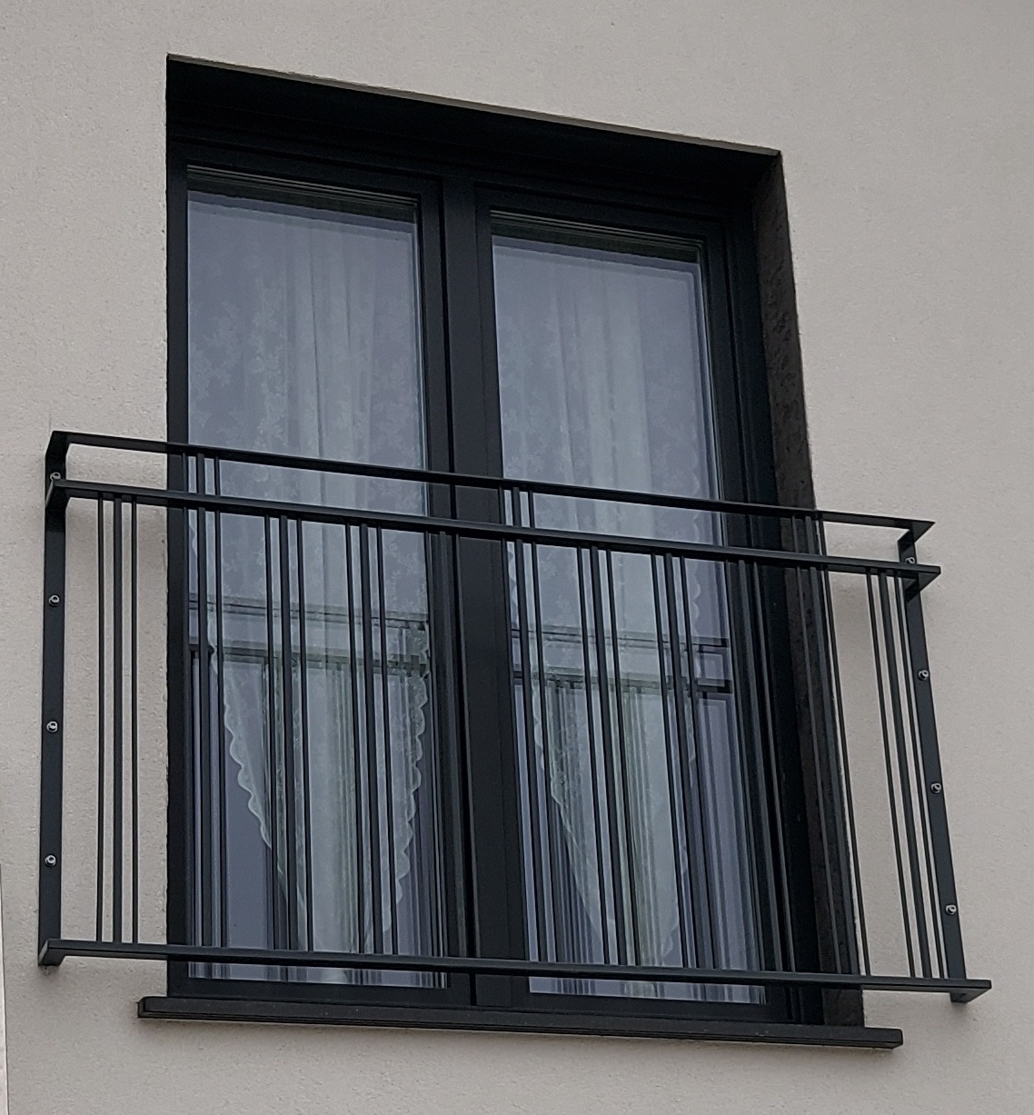 Czarna, metalowa balustrada okienna z pionowymi prętami, zamontowana na elewacji budynku. Widok na okno z firanką w tle, z zewnątrz.