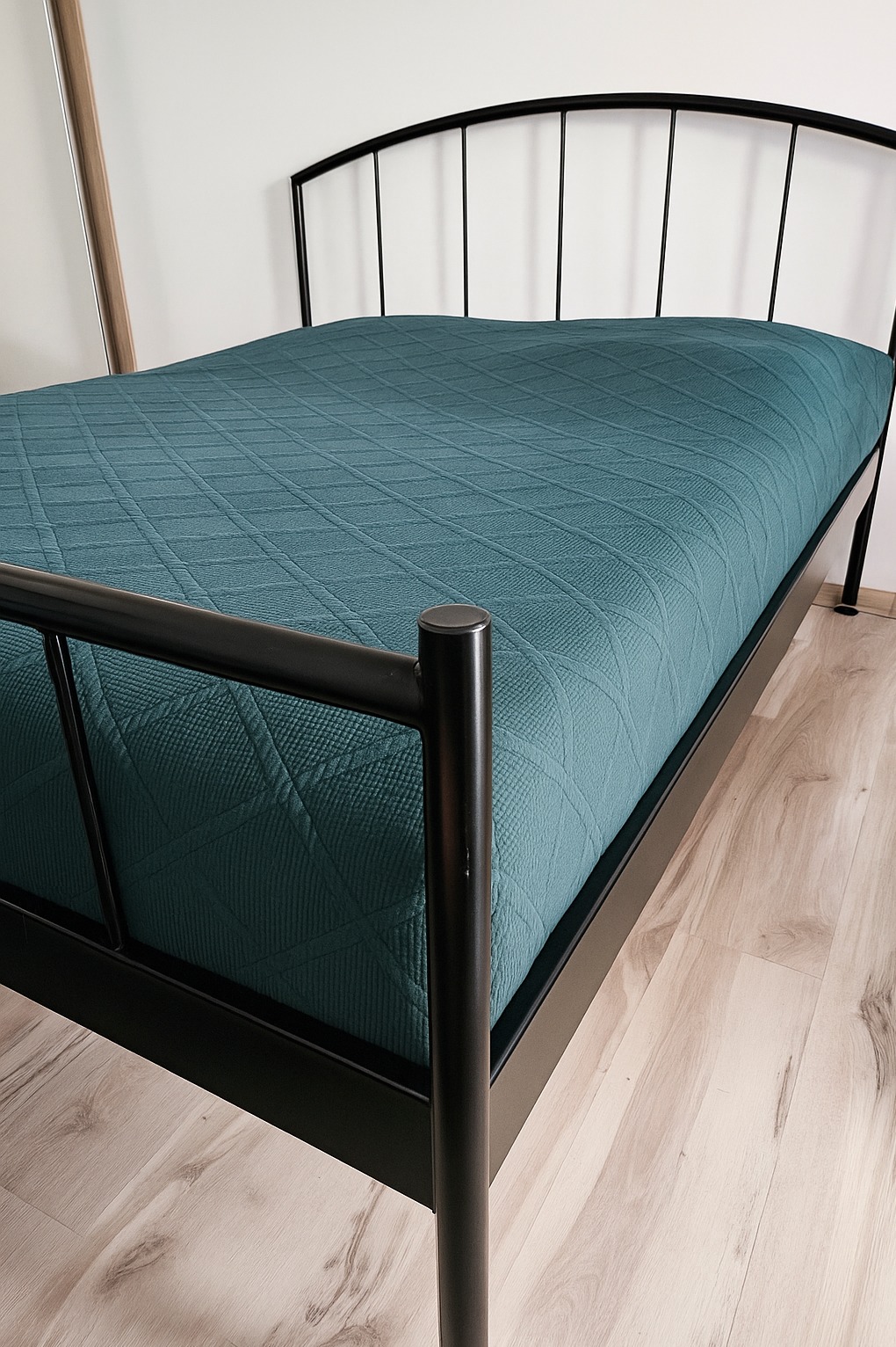 Czarne metalowe łóżko z turkusową narzutą o geometrycznym wzorze, na jasnej podłodze. Widoczne odbicie w lustrze i fragment białej ściany. Minimalistyczny design.