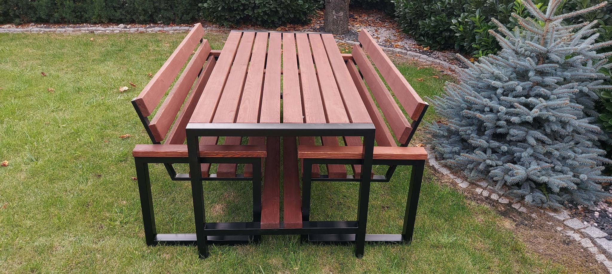 Stół piknikowy z drewna i metalu na trawie, otoczony zielenią. Stół z ławkami w nowoczesnym stylu, z czarną metalową ramą i drewnianymi elementami w ciepłym odcieniu brązu.