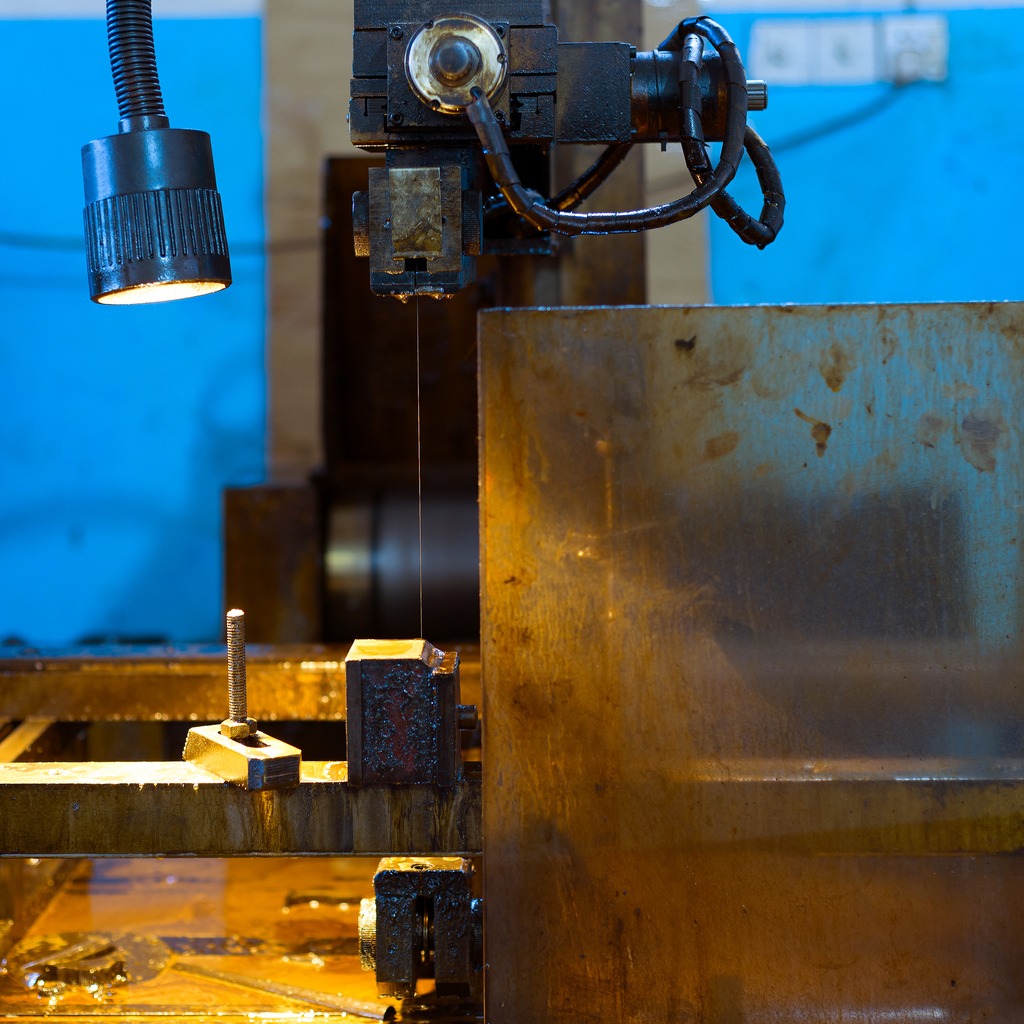 Elektroerozyjna obróbka drutowa CNC: zbliżenie na proces cięcia metalu. Widoczny drut tnący, chłodziwo i obrabiany element. Oświetlenie robocze.