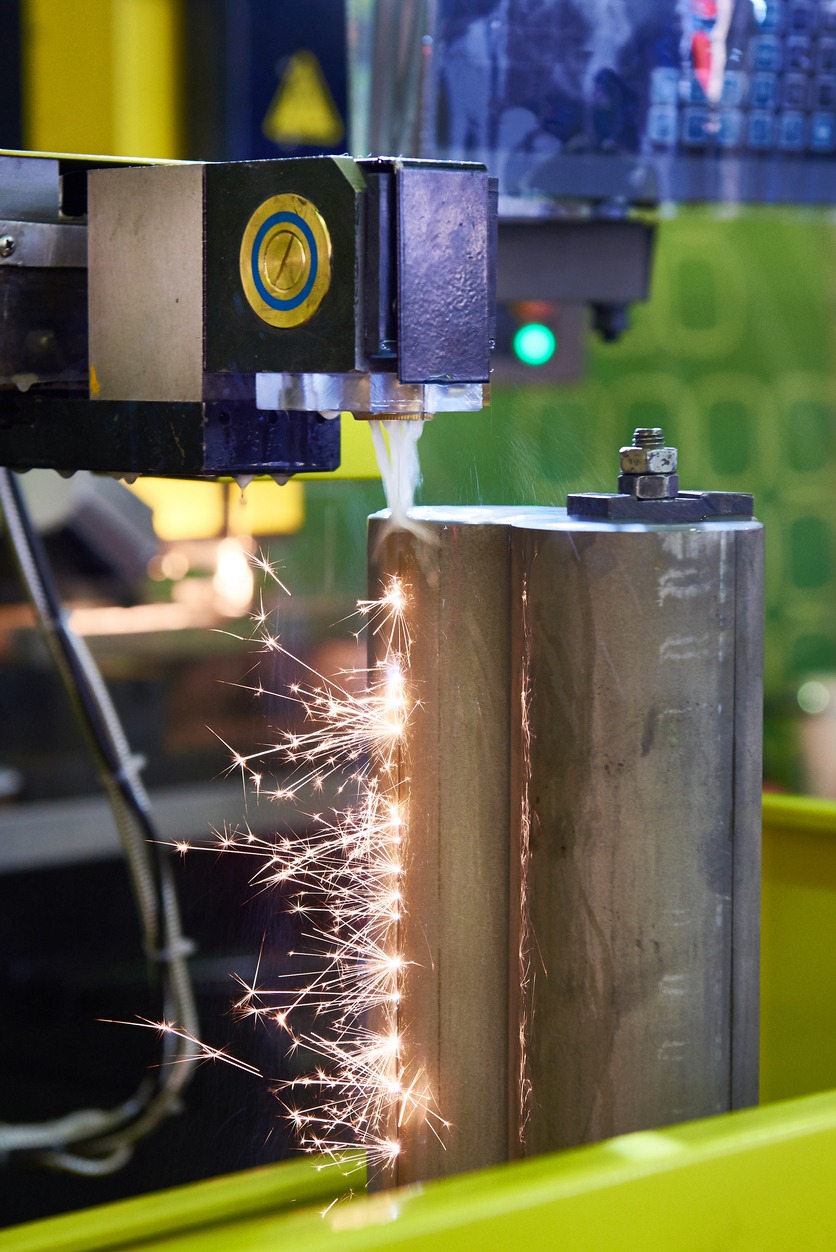 Elektroerozyjna obróbka metalu: iskrzenie podczas cięcia pionowej, metalowej kolumny za pomocą precyzyjnej maszyny. Chłodziwo spływa po obrabianym elemencie.