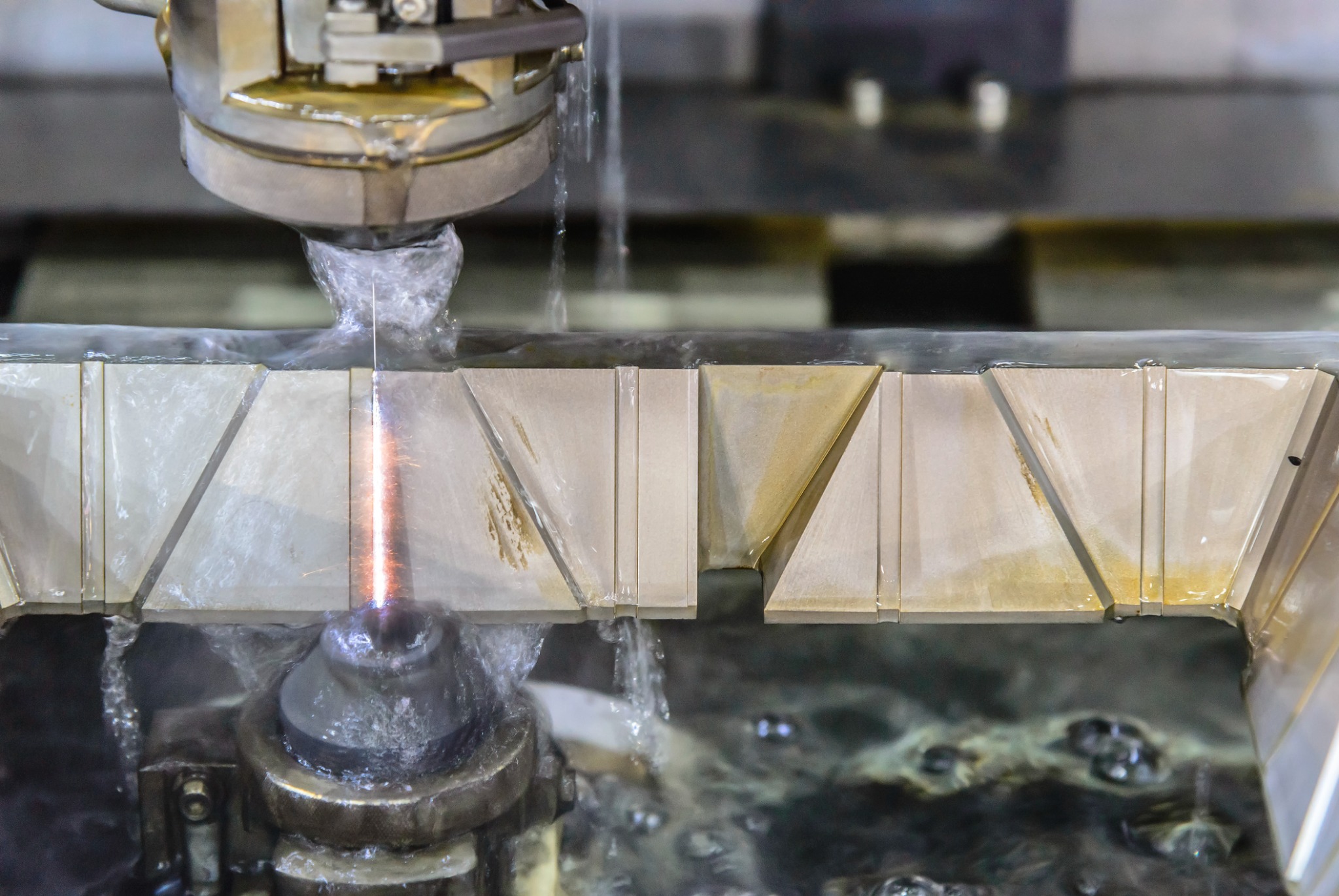 Elektroerozyjna obróbka metalu: drut tnący zanurzony w cieczy dielektrycznej, precyzyjnie wycinający wzór w metalowym elemencie o geometrycznych kształtach.