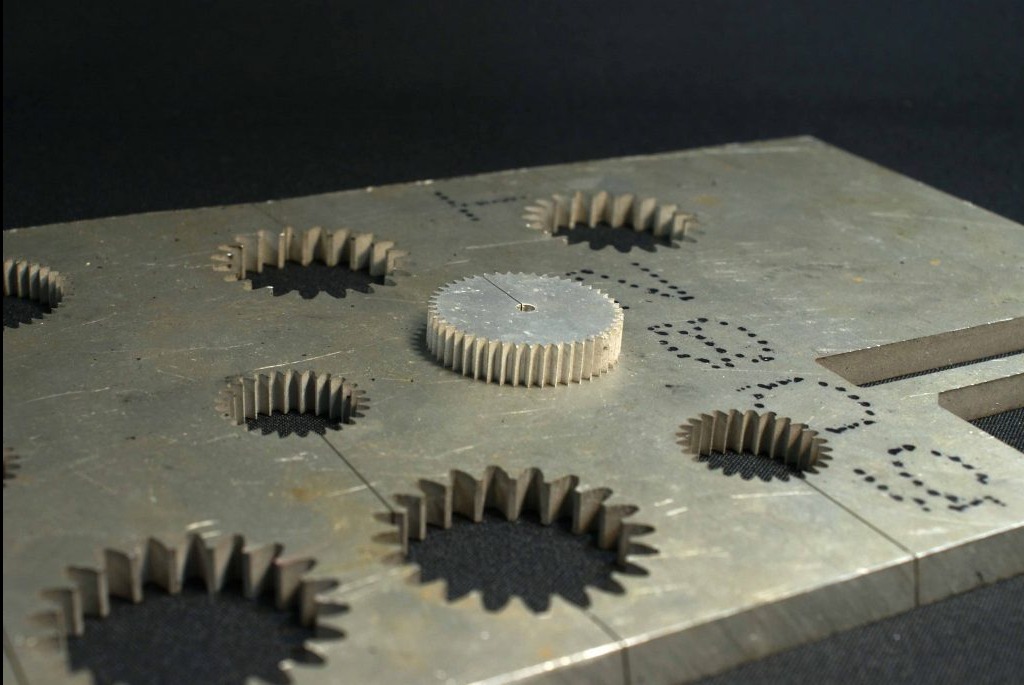 Metalowa płyta z wyciętymi otworami na koła zębate i jednym kołem umieszczonym na płycie. Widoczne ślady obróbki i oznaczenia punktowe na powierzchni.