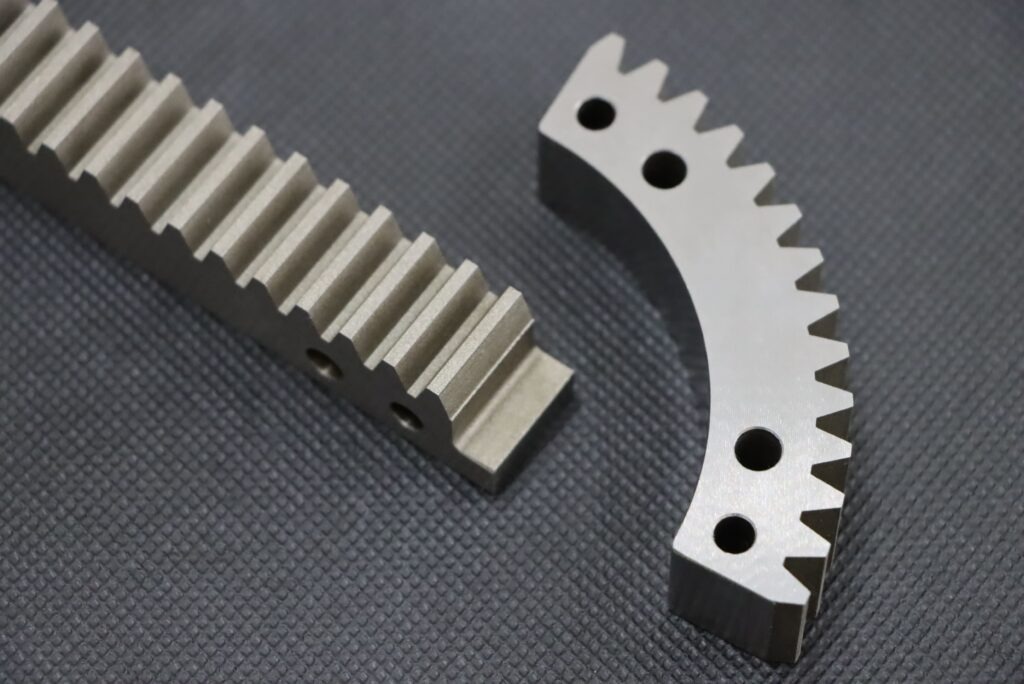 Precyzyjnie wykonane, metalowe listwy zębate: prosta i łukowa, z widocznymi otworami montażowymi, leżą na szarym tle. Detal obróbki.