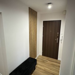 Maciej Dec - Nowoczesny przedpokój z drewnianymi lamelami na ścianie, czarną ławką i brązowymi płytkami podłogowymi. Minimalistyczne oświetlenie sufitowe.