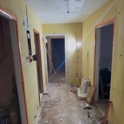 Maciej Dec - Korytarz w trakcie prac remontowych: ściany przygotowane do malowania, widoczne narzędzia, worki z zaprawą, lampa oświetleniowa. Podłoga częściowo zakryta resztkami materiałów.
