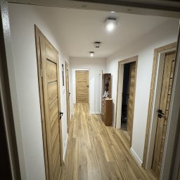 Maciej Dec - Długi korytarz z drewnianą podłogą i białymi ścianami. Widoczne drzwi z drewnianymi okleinami i czarnymi klamkami. Oświetlenie sufitowe punktowe.