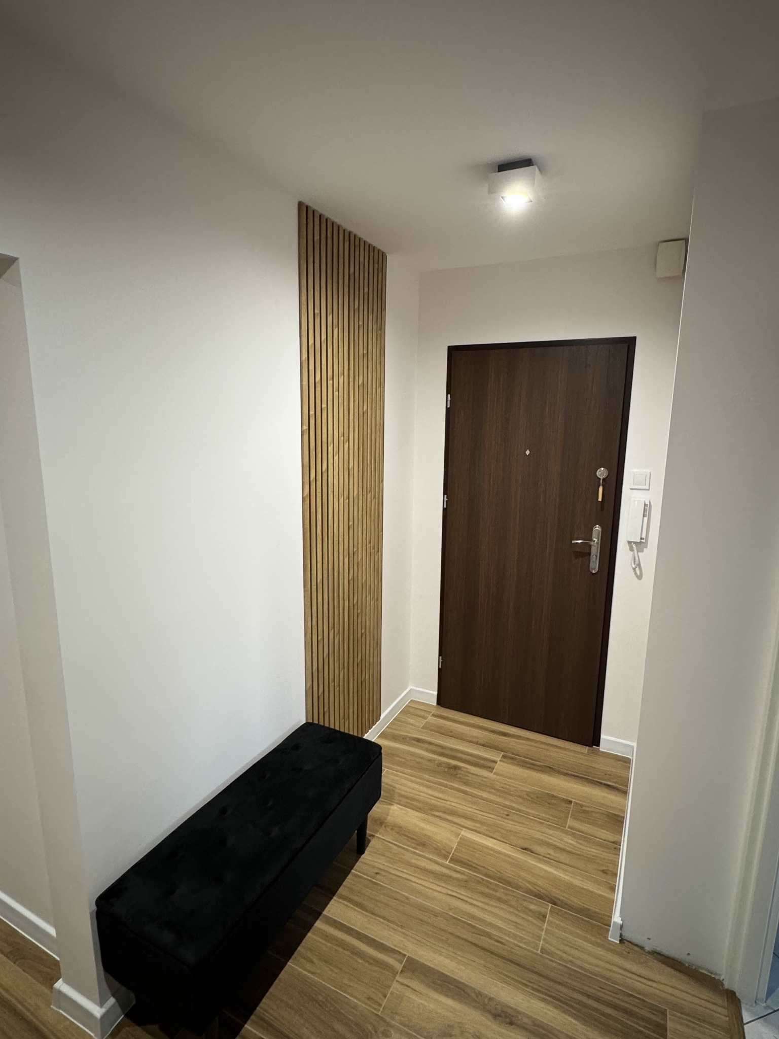 Nowoczesny przedpokój z drewnianymi lamelami na ścianie, czarną ławką i brązowymi płytkami podłogowymi. Minimalistyczne oświetlenie sufitowe.