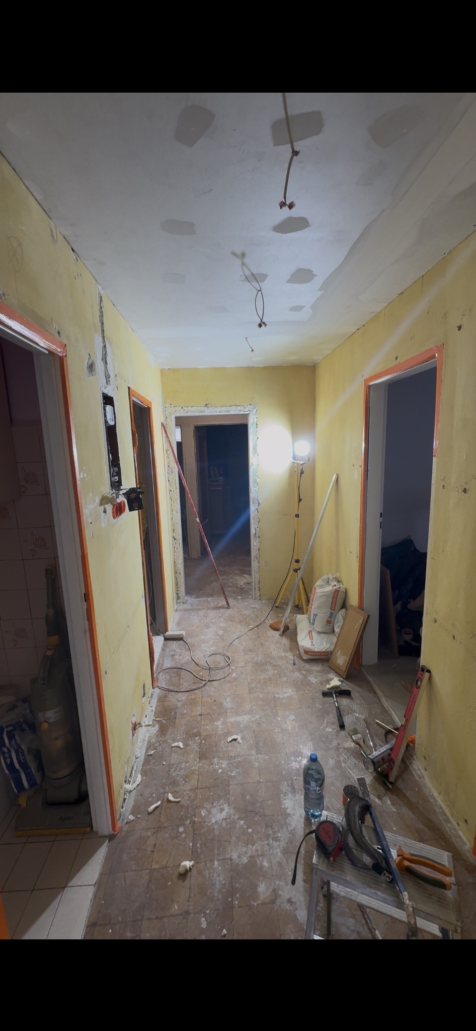Korytarz w trakcie prac remontowych: ściany przygotowane do malowania, widoczne narzędzia, worki z zaprawą, lampa oświetleniowa. Podłoga częściowo zakryta resztkami materiałów.