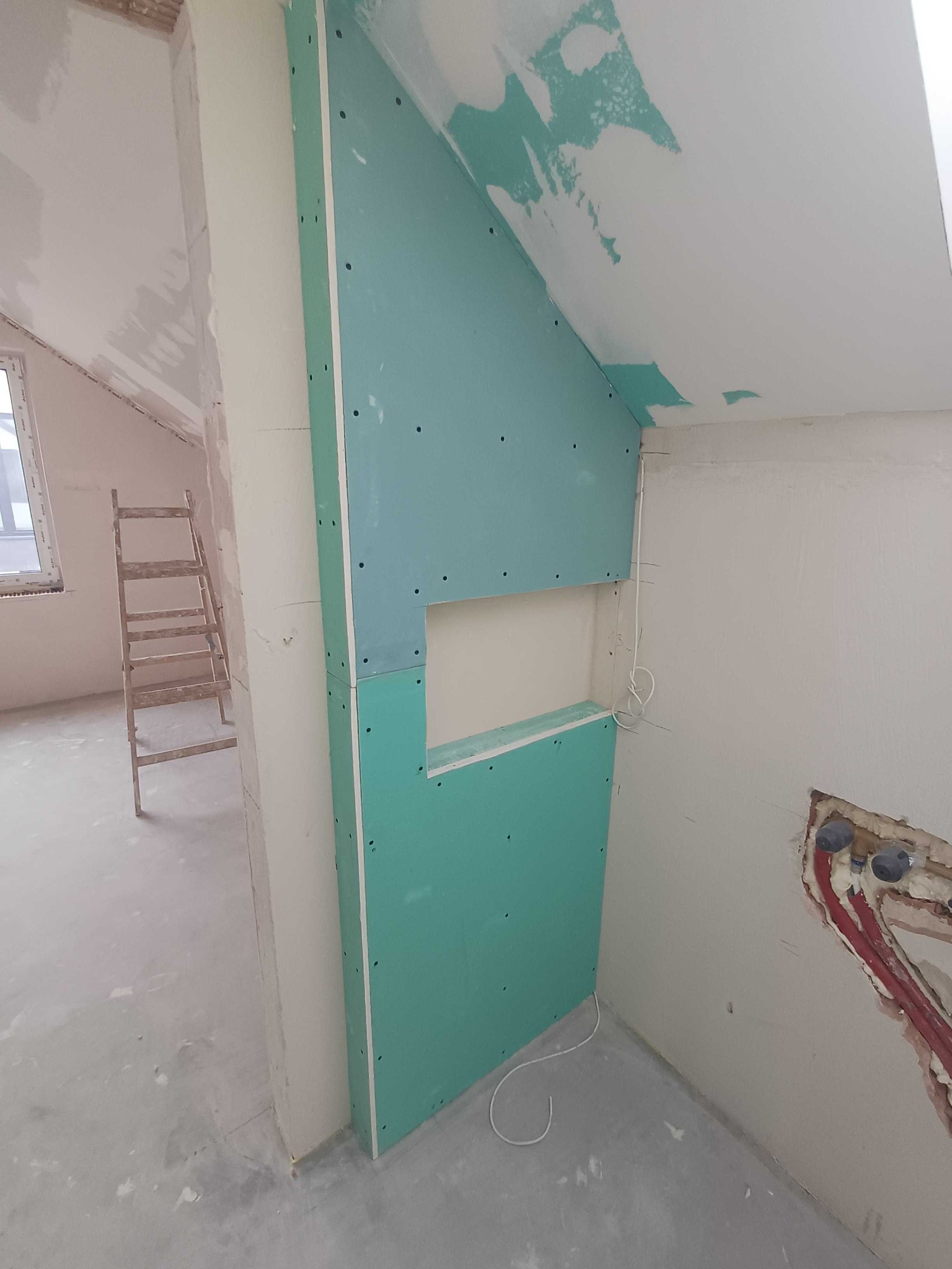 Montaż zielonej płyty GK z otworem na ścianie pod skosem. Widoczne instalacje, drabina w tle. Surowy stan wykończenia wnętrza, przygotowanie do dalszych prac.